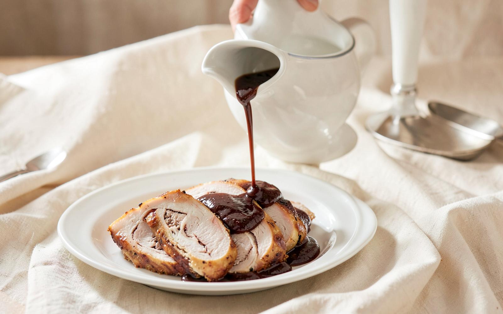 Glossy turkey gravy poured from a white ceramic pitcher over sliced turkey roulade on a cream plate